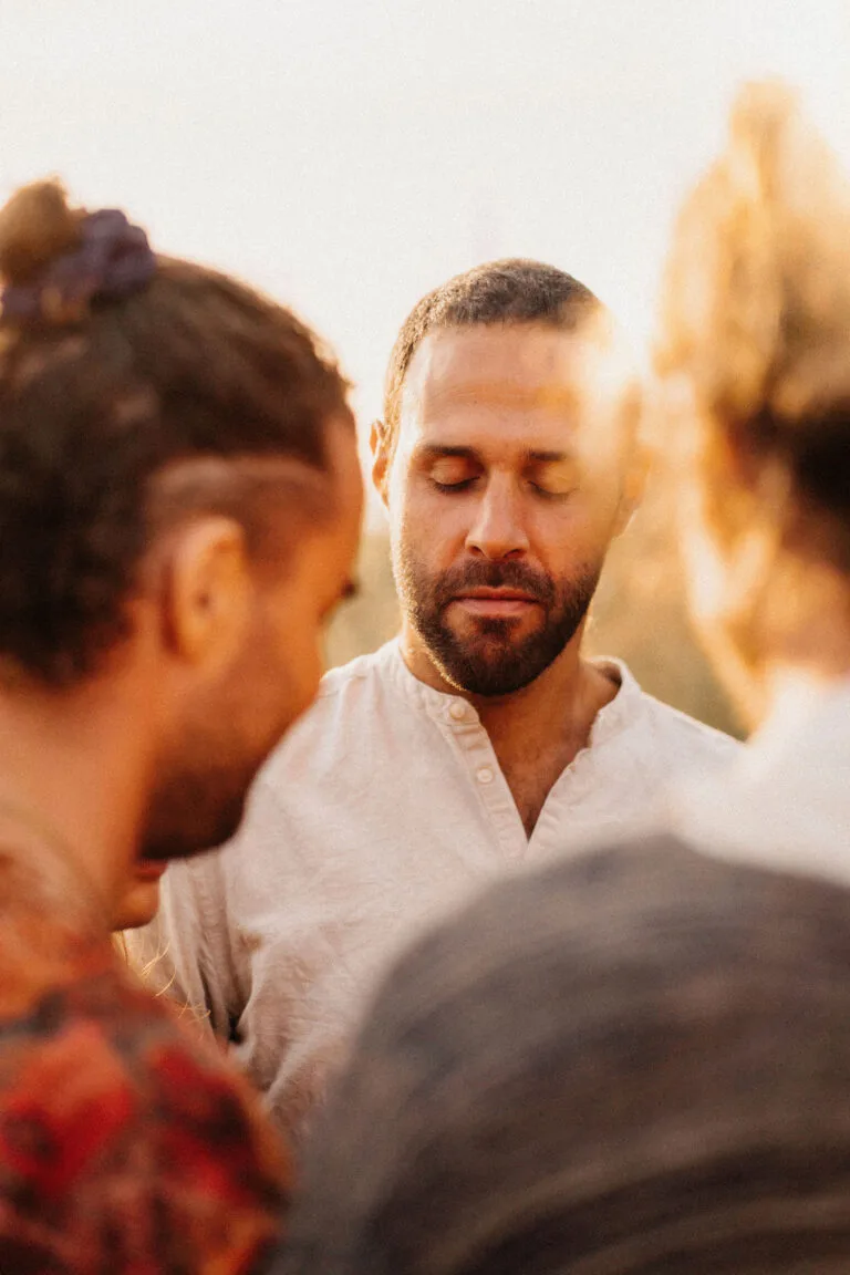 Sound healer facing the camera with eyes closed, surrounded by a circle of participants, attentively listening to someone share their experience after a sound healing exercise.